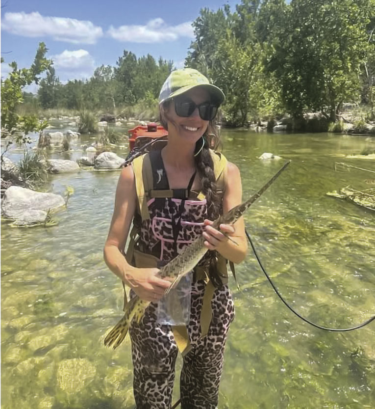 Woman smiling in river, holding a fish, wearing a green cap, sunglasses, and patterned waders. Bright, sunny day with trees and rocks around.