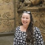 Smiling woman with long hair in a patterned dress stands in front of ornate, carved stone wall. The setting conveys a historic or artistic theme.