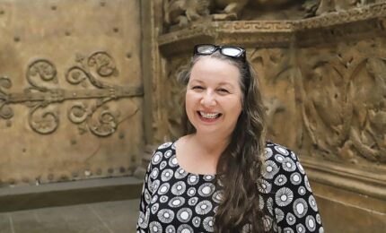 Smiling woman with long hair in a patterned dress stands in front of ornate, carved stone wall. The setting conveys a historic or artistic theme.