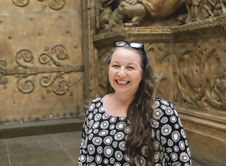 Smiling woman with long hair in a patterned dress stands in front of ornate, carved stone wall. The setting conveys a historic or artistic theme.