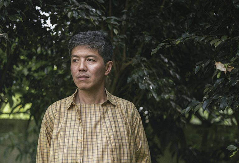 Middle-aged man in a plaid shirt stands outdoors, gazing thoughtfully to the right. Lush green foliage forms the background, creating a calm atmosphere.