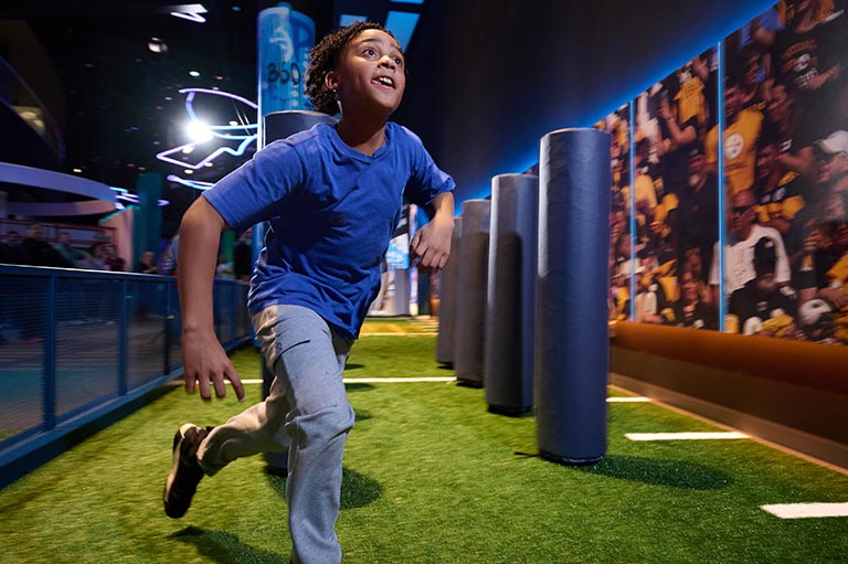 A child in a blue shirt runs enthusiastically through an interactive exhibit with padded columns on a synthetic field, evoking energy and excitement.