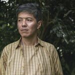 Middle-aged man in a plaid shirt stands outdoors, gazing thoughtfully to the right. Lush green foliage forms the background, creating a calm atmosphere.