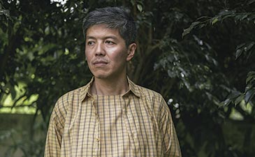 Middle-aged man in a plaid shirt stands outdoors, gazing thoughtfully to the right. Lush green foliage forms the background, creating a calm atmosphere.