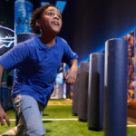 A child in a blue shirt runs enthusiastically through an interactive exhibit with padded columns on a synthetic field, evoking energy and excitement.
