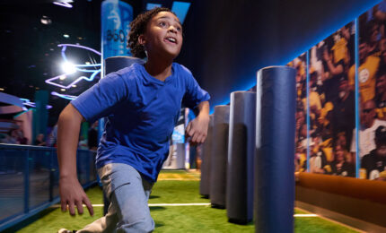 A child in a blue shirt runs enthusiastically through an interactive exhibit with padded columns on a synthetic field, evoking energy and excitement.
