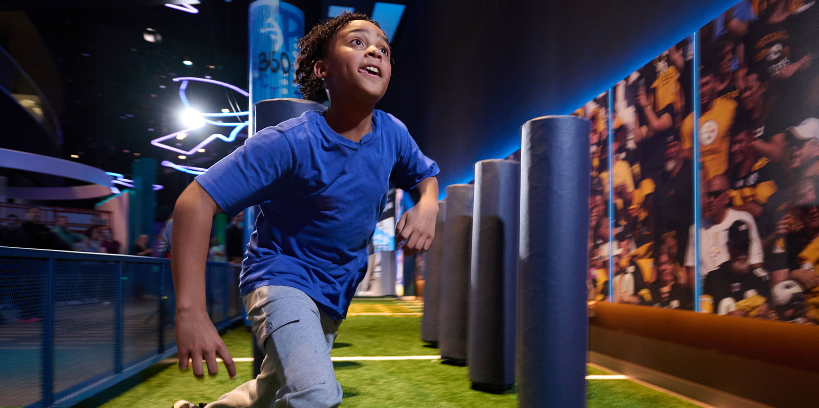 A child in a blue shirt runs enthusiastically through an interactive exhibit with padded columns on a synthetic field, evoking energy and excitement.