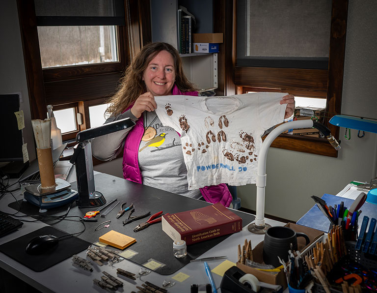 A woman smiles while holding a T-shirt with brown animal footprints at a desk cluttered with books, tools, and office supplies, conveying curiosity and enthusiasm.
