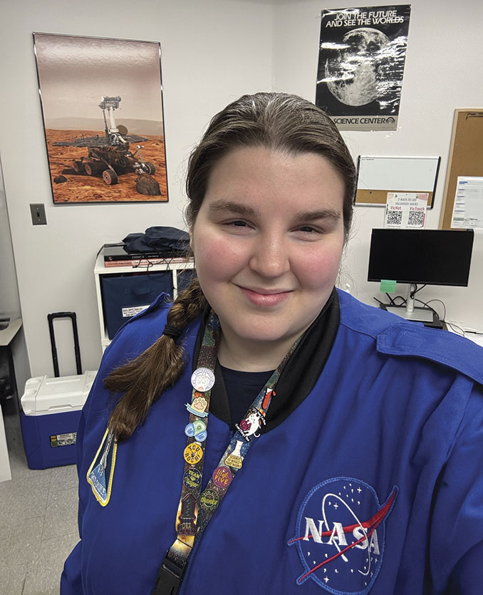 Person in a blue NASA jacket smiles in an office. Background features a Mars rover poster, a computer, and various space-themed décor. Warm, enthusiastic tone.