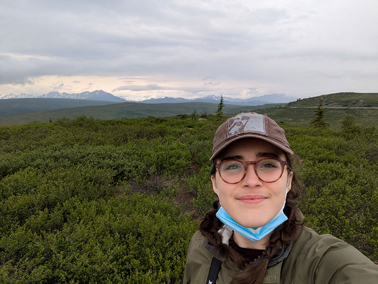 A woman with glasses and a brown cap smiles in a lush green landscape under a cloudy sky, with distant mountains. The scene conveys a serene and cheerful mood.