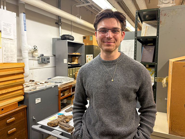 A person in a gray sweater stands smiling in a cluttered research lab. Shelves of books and fossils surround them, conveying a scholarly atmosphere.