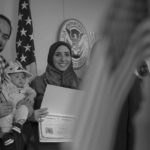 A family posing at anaturalization ceremony tthe U.S. Citizenship and Immigration Services office man is holding an american flag and a baby standing in front of a flag