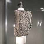 A textured gray rock sculpture displayed on a white pedestal, showcasing its unique natural patterns under soft lighting.