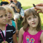 A person holds a small bird, while several curious children gather around, intrigued by the interaction in a lush outdoor setting.