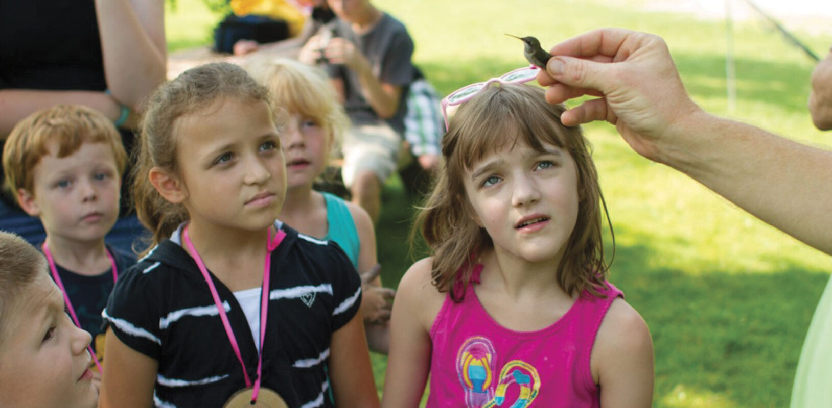 A person holds a small bird, while several curious children gather around, intrigued by the interaction in a lush outdoor setting.