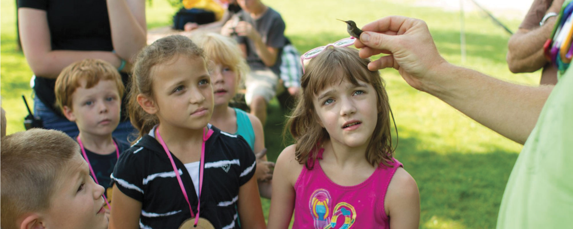 A person holds a small bird, while several curious children gather around, intrigued by the interaction in a lush outdoor setting.