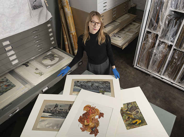 A person wearing blue gloves stands over framed artwork in a storage area filled with art supplies and organized drawers.