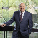 A man in a dark suit and patterned tie leans against a glass railing, with green trees visible in the background.