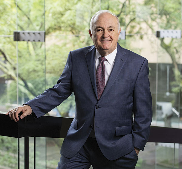 A man in a dark suit and patterned tie leans against a glass railing, with green trees visible in the background.