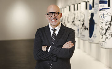 A man in a suit stands confidently with arms crossed in an art gallery, featuring colorful artworks on white elongated panels.