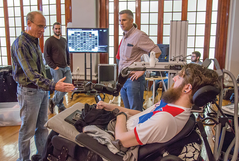 three engineers gathered around a man in a a wheelchair testing a robotic hand