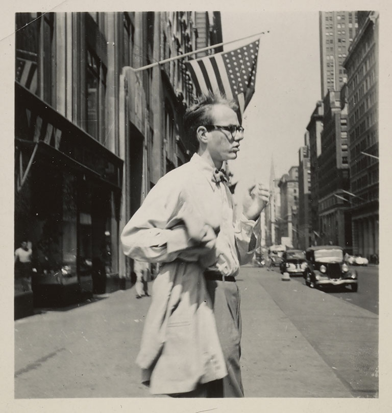 Young andy Warhol crossing the street in 1950's New York City dressed in a dress shirt, bow tie, and carrying a sport coat.