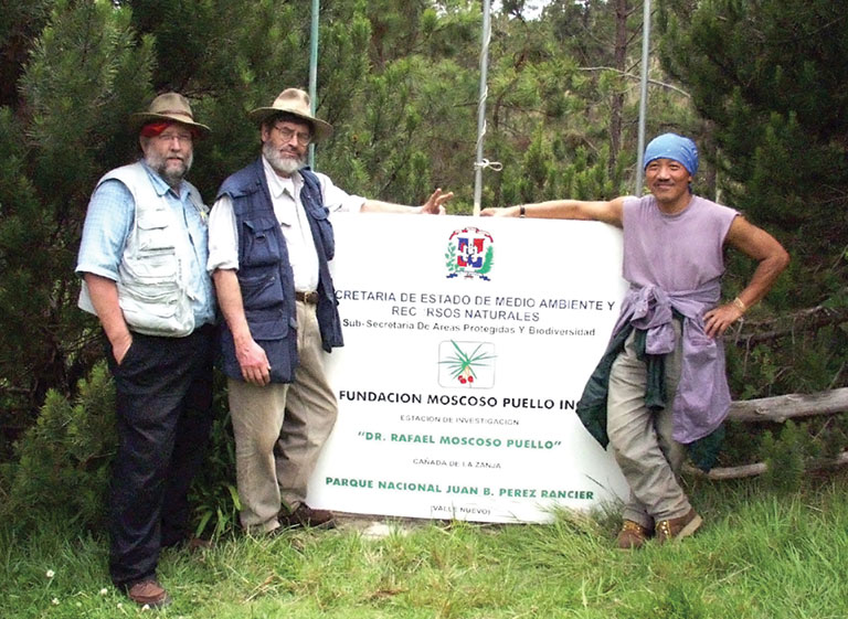 portrait of John Rawlins, Bob Davidson, and Chen Young standing together in front of a sign in the Dominican Republic