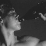 A still from a black and white Warhol movie of a bare chested young man, drinking out of a bottle