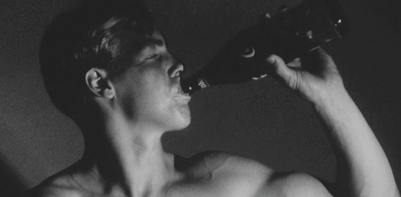 A still from a black and white Warhol movie of a bare chested young man, drinking out of a bottle
