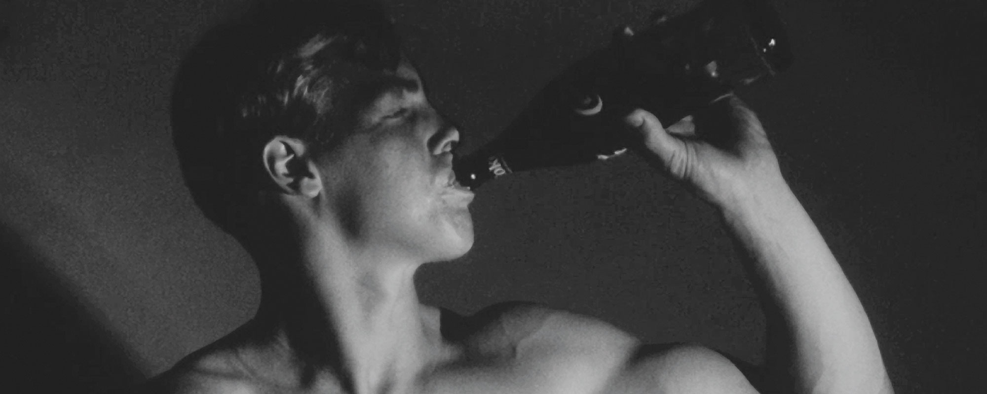 A still from a black and white Warhol movie of a bare chested young man, drinking out of a bottle