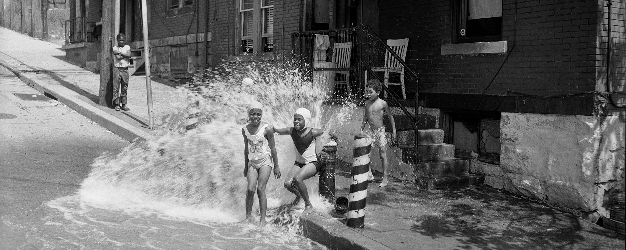 A black and white photo from the 1940's showing a group of children playing in a fire hydrant on the street.