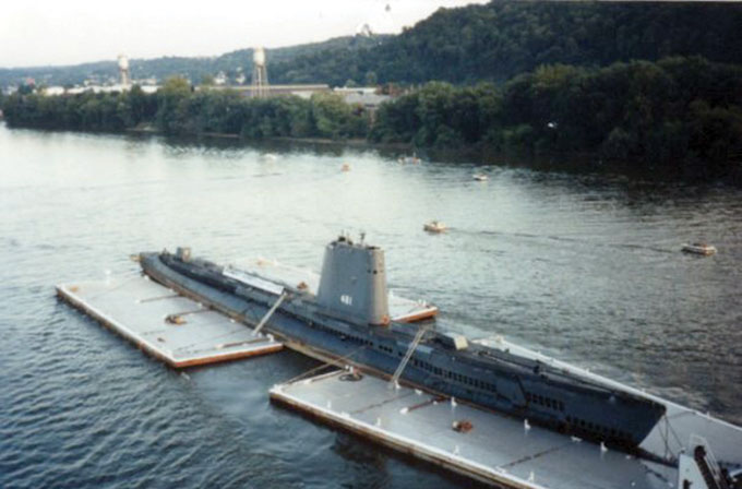 A submarine being transported up a river towed by barges