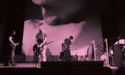 Black and white photo of a band on stage playing music with a film of ttwo people kissing projected behind them