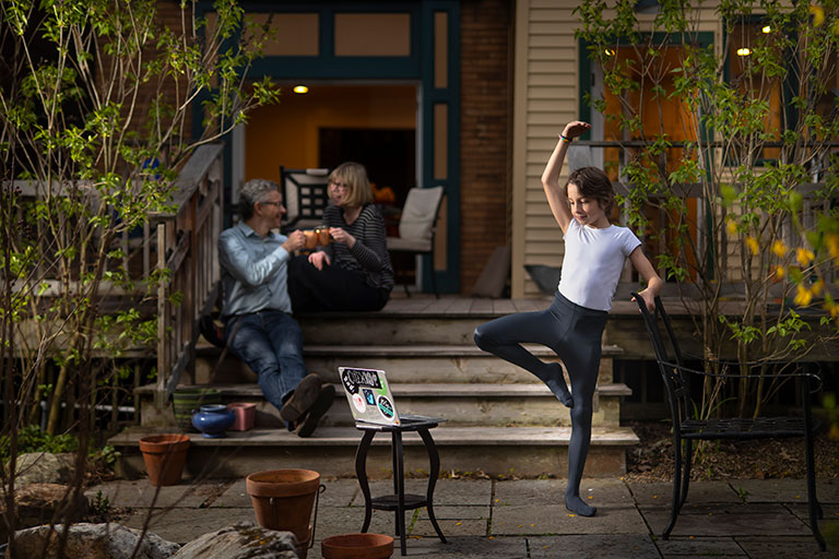 A young boy practicing ballet outside of his home with his parents in the background.