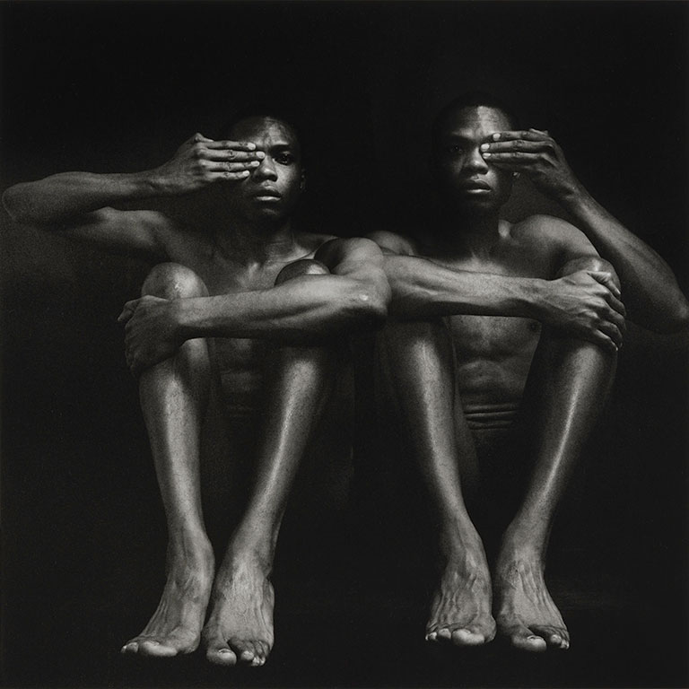 Photo of 2 men sitting side by side each covering one of their eyes.