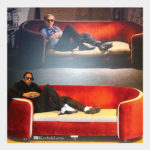A photo of a man on a red couch sittign in front of an image of Warhol on a similar couch