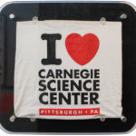 A t-shirt with the words I heart Carnegie SCience center, framed in a display case.