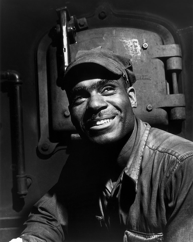 A black and white portrait of a man smiling at teh camera wearing work clothes.