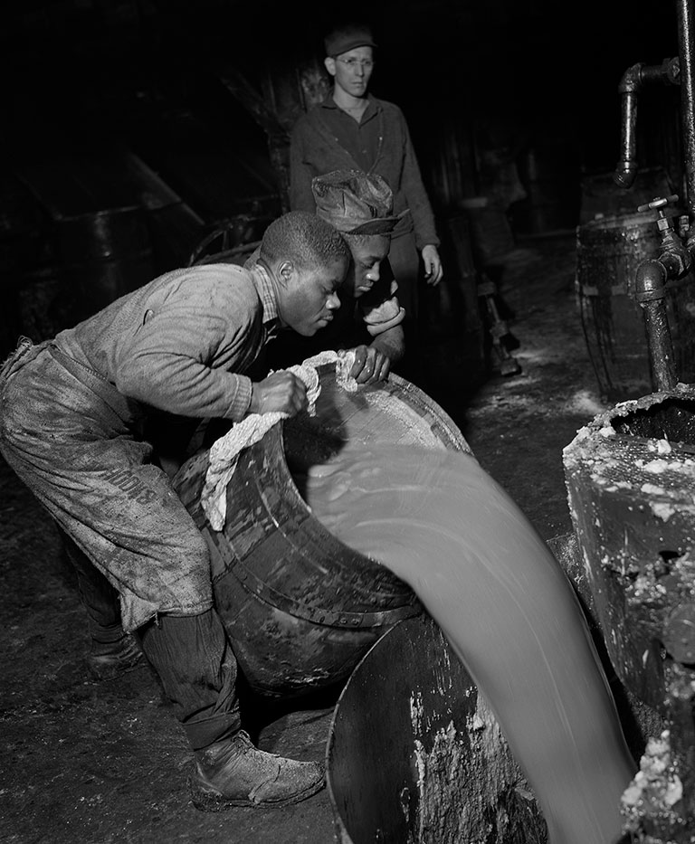 A black and white photo of 2 men pouring grease from a large barrel. .