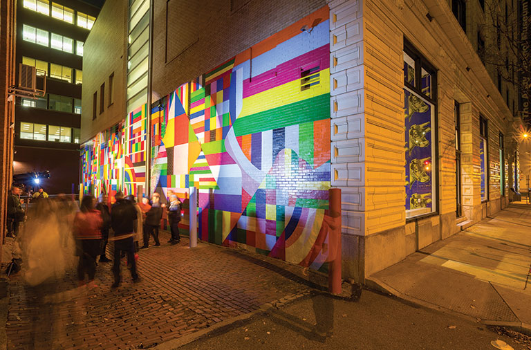 A photo of an alley way witha colorful mural painted on one side. Blurred images of people are scattered in the image.