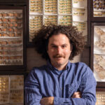 A man with curly hair surrounded by cases of butterfly and moth specimens.