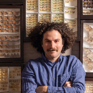 A man with curly hair surrounded by cases of butterfly and moth specimens.