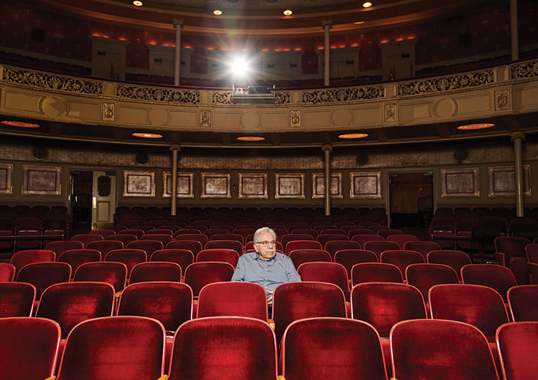A man sitting alone in a theater in red velvet seats.