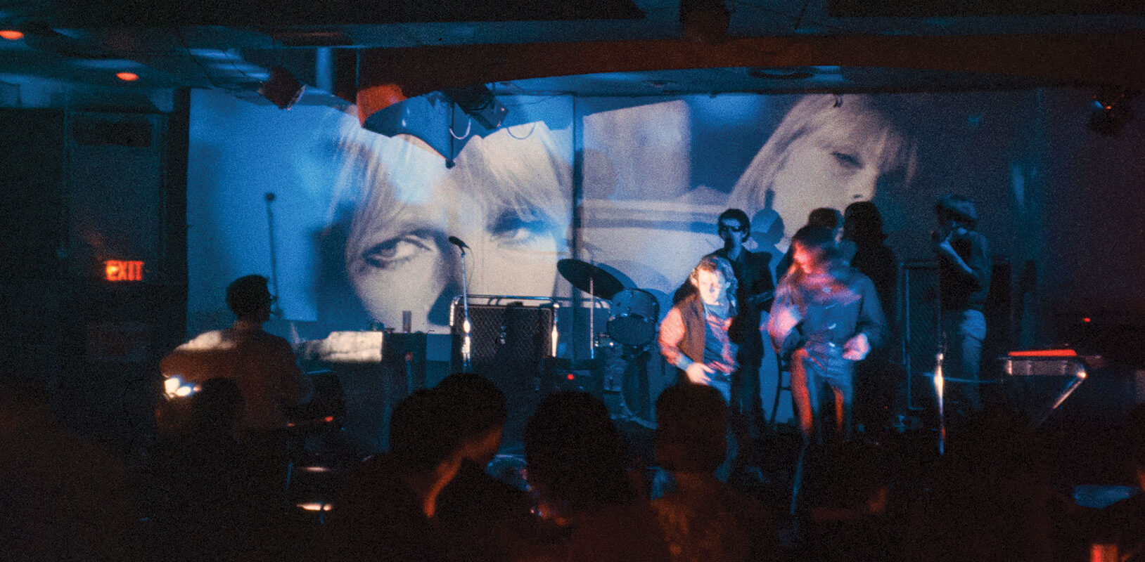 Color photograph of three of the members of the Velvet Underground performing on a stage as images are being projected onto the walls behind them.