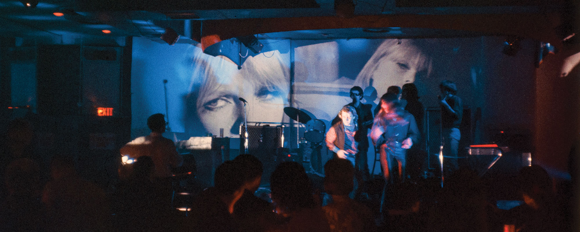 Color photograph of three of the members of the Velvet Underground performing on a stage as images are being projected onto the walls behind them.