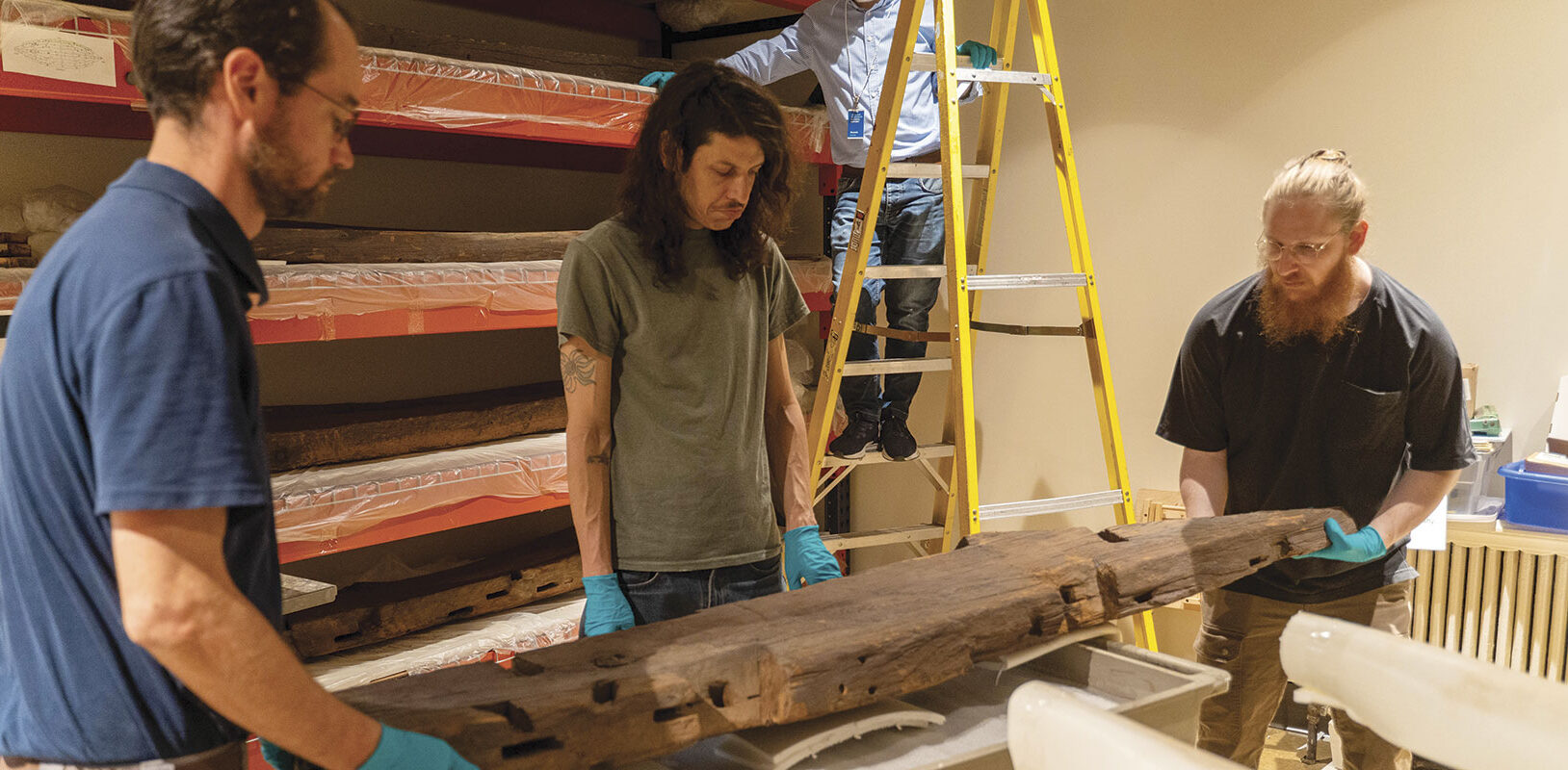 Three museum workers moving a large wooden beam.