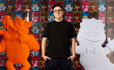 A portrait of the artist, Kaws standing on front of some of his work.