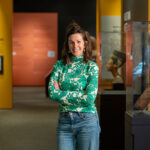 A person in a green floral top stands with arms crossed in a museum, with colorful exhibit walls and artifacts displayed nearby.