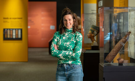 A person in a green floral top stands with arms crossed in a museum, with colorful exhibit walls and artifacts displayed nearby.