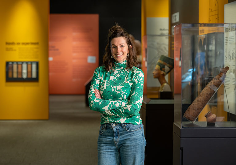 A person in a green floral top stands with arms crossed in a museum, with colorful exhibit walls and artifacts displayed nearby.